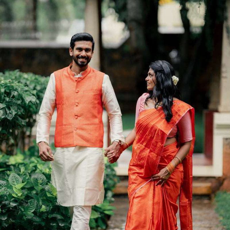 A couple walking hand in hand through a garden, he in an orange waistcoat over a cream kurta, she in an orange silk saree